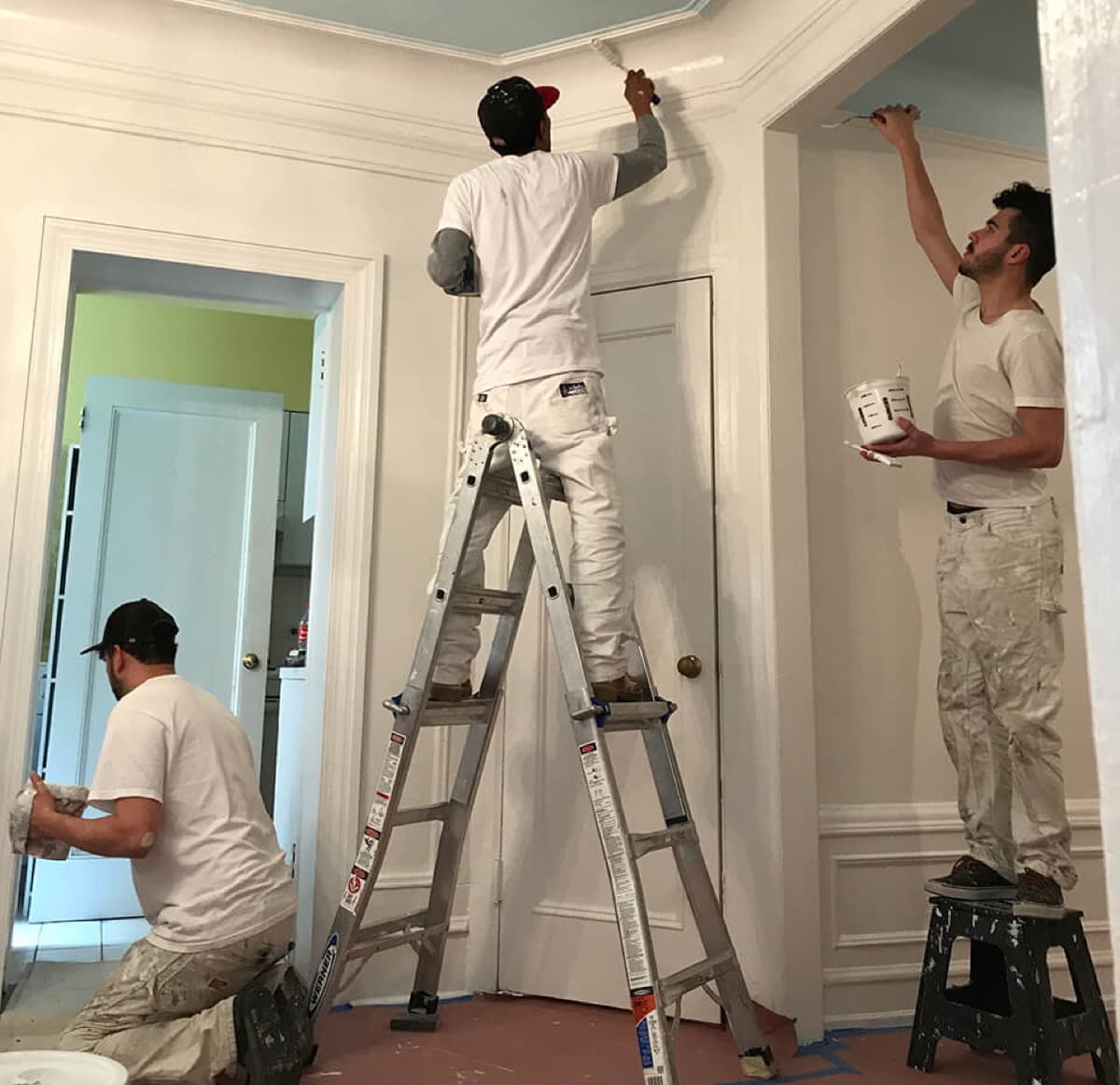 Two painters at work in a white room, one on a ladder painting the door trim