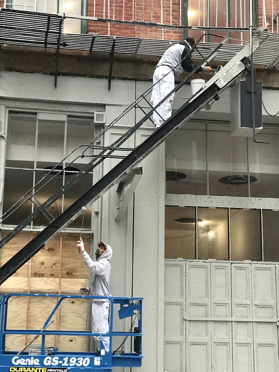 Painter on a ladder beside a scissor lift at a building exterior